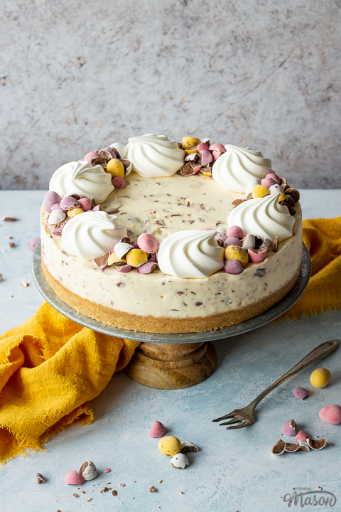 A whole no bake Mini Egg cheesecake on a cake stand topped with mini meringue nests and chopped Mini Eggs. There is a fork, chopped mini eggs and a yellow linen napkin in the background, all set on a light blue backdrop.