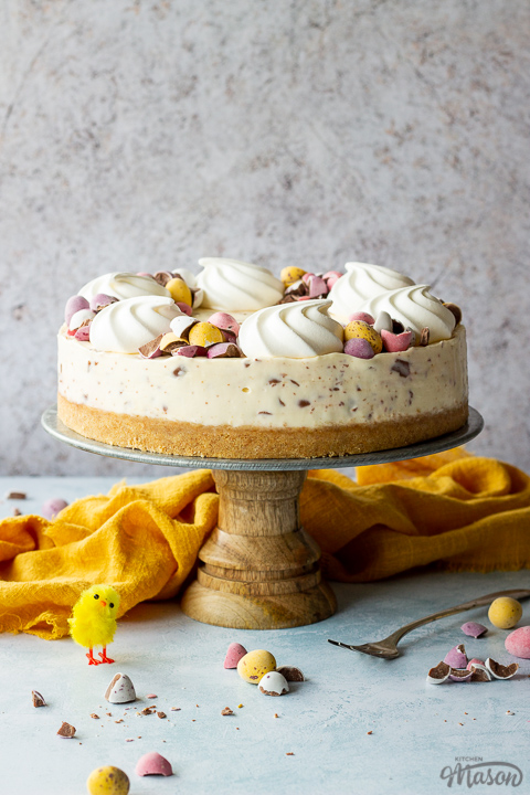 A whole no bake Mini Egg cheesecake on a cake stand topped with mini meringue nests and chopped Mini Eggs. There is a fork, chopped mini eggs and a yellow linen napkin in the background, all set on a light blue backdrop.