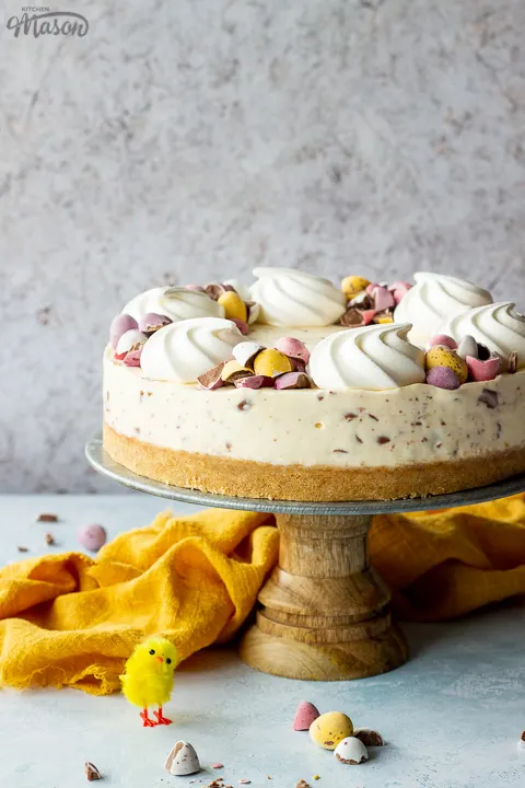 A whole no bake Mini Egg cheesecake on a cake stand topped with mini meringue nests and chopped Mini Eggs. There is chopped mini eggs and a yellow linen napkin in the background, all set on a light blue backdrop.