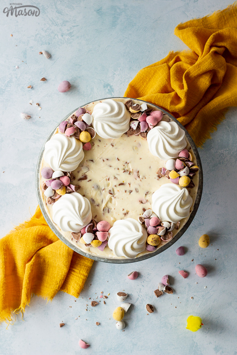 Birds eye view of a no bake Mini Egg cheesecake topped with mini meringue nests and chopped Mini Eggs on a cake stand set on a light blue backdrop. There are chopped Mini eggs and a yellow linen napkin in the background.
