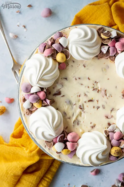 Birds eye view of a no bake Mini Egg cheesecake topped with mini meringue nests and chopped Mini Eggs on a cake stand set on a light blue backdrop. There are chopped Mini eggs. a yellow linen napkin and a fork in the background.