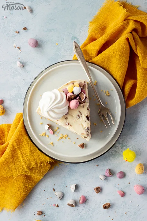 A slice of no bake Mini Egg cheesecake topped with a mini meringue nest and chopped Mini Eggs on two stacked plates with a fork on the side. There are chopped mini eggs and a yellow linen napkin set over a light blue background.