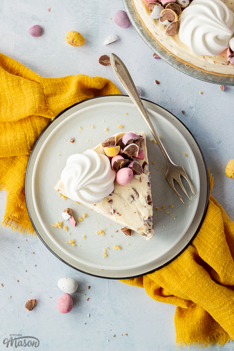A slice of no bake Mini Egg cheesecake topped with a mini meringue nest and chopped Mini Eggs on two stacked plates with a fork on the side. There are chopped mini eggs, a yellow linen napkin and the remaining cheesecake on a cake stand set over a light blue background.