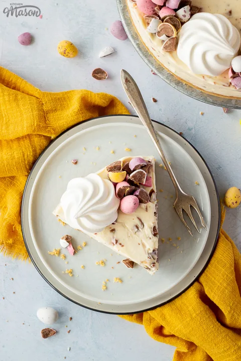A slice of no bake Mini Egg cheesecake topped with a mini meringue nest and chopped Mini Eggs on two stacked plates with a fork on the side. There are chopped mini eggs, a yellow linen napkin and the remaining cheesecake on a cake stand set over a light blue background.
