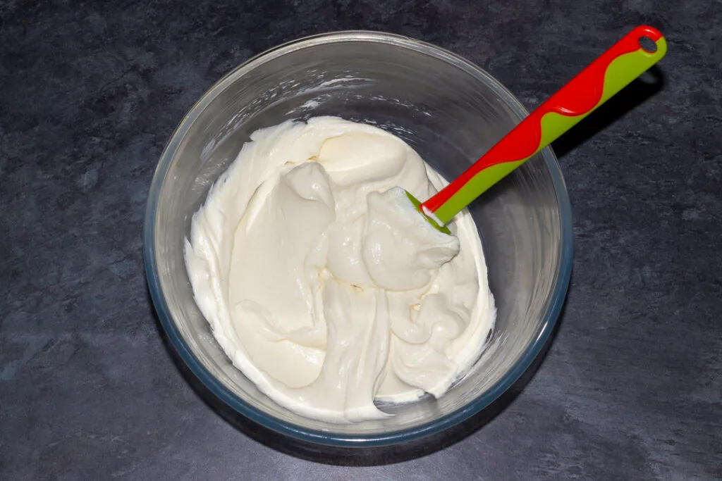 Cream cheese and caster sugar beaten together in a glass bowl with a spatula on a kitchen worktop