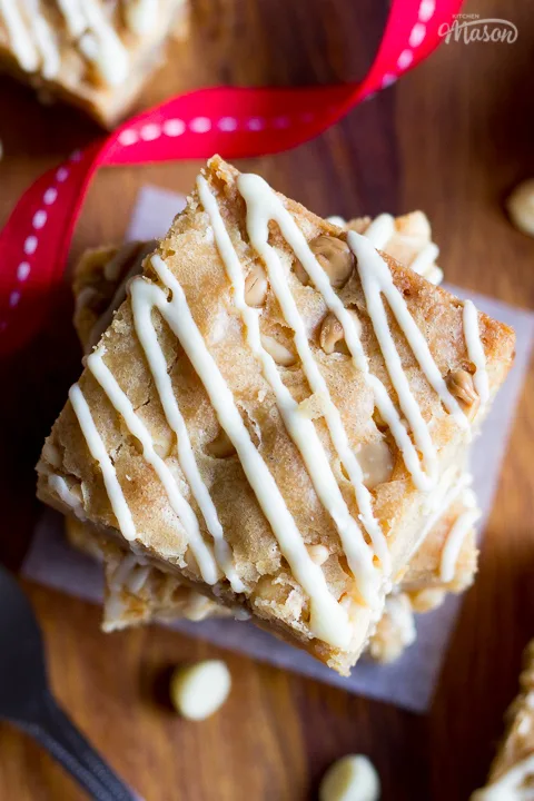 Flat lay view of stacked and individual white chocolate blondies on a dark wood worktop. There are white chocolate chips scattered around, a fork and a red ribbon swirling through the middle.