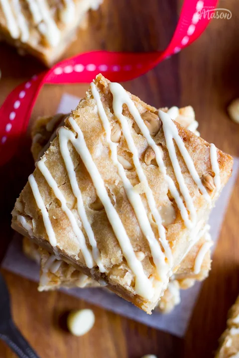 Flat lay view of stacked and individual white chocolate blondies on a dark wood worktop. There are white chocolate chips scattered around, a fork and a red ribbon swirling through the middle.