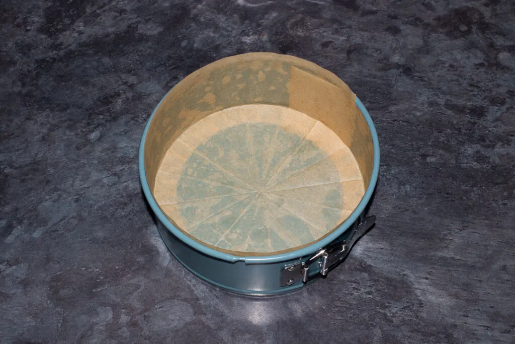 A springform pan lined with baking paper on the base and sides on a kitchen worktop.