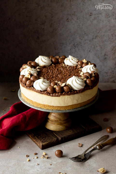 A no bake Malteser cheesecake on a cake stand. With crushed Maltesers scattered around, a red linen napkin and 2 forks. Set on a light neutral backdrop.