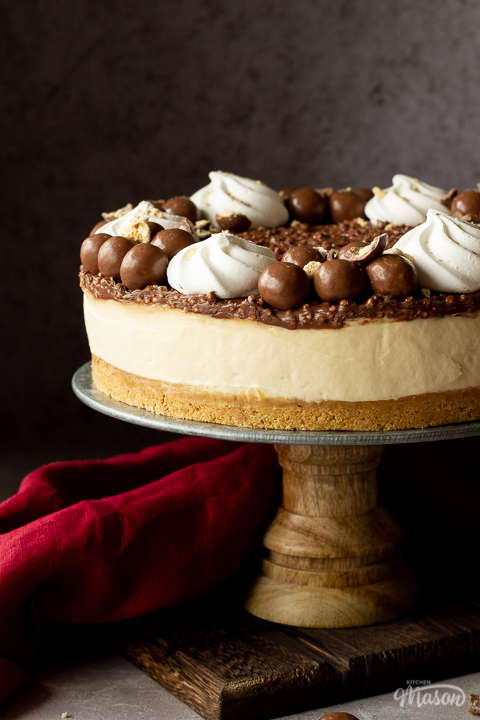 Side view of a no bake Malteser cheesecake on a cake stand. With a red linen napkin in the background, set on a light neutral backdrop.