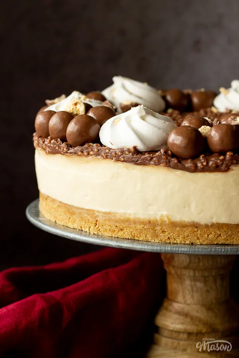 Close up side view of a no bake Malteser cheesecake on a cake stand. With a red linen napkin in the background, set on a light neutral backdrop.
