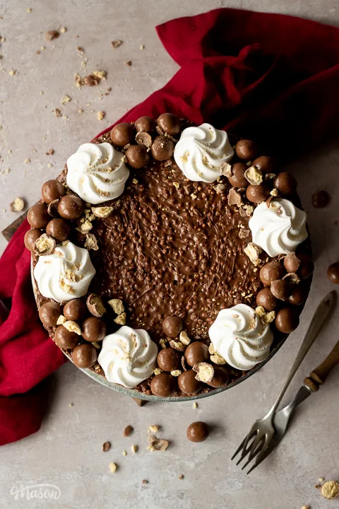Birds eye view of a no bake Malteser cheesecake on a cake stand with crushed Maltesers scattered around, 2 forks and a red linen napkin in the background. Set on a light neutral backdrop.