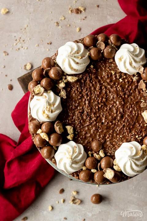 Close up birds eye view of a no bake Malteser cheesecake on a cake stand with crushed Maltesers scattered around, 2 forks and a red linen napkin in the background. Set on a light neutral backdrop.
