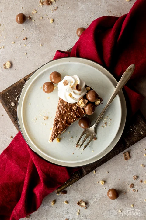 Birds eye view of a slice of no bake Malteser cheesecake on 2 stacked plates with a fork. There are crushed Maltesers scattered around and a red linen napkin in the background. Set on a light neutral backdrop.