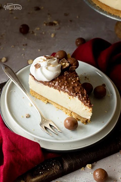 Angled view of a slice of no bake Maltesers cheesecake set on 2 stacked plates with a fork. There are crushed Maltesers scattered around, a red linen napkin and the remaining cheesecake on a cake stand in the background. Set on a light neutral backdrop.