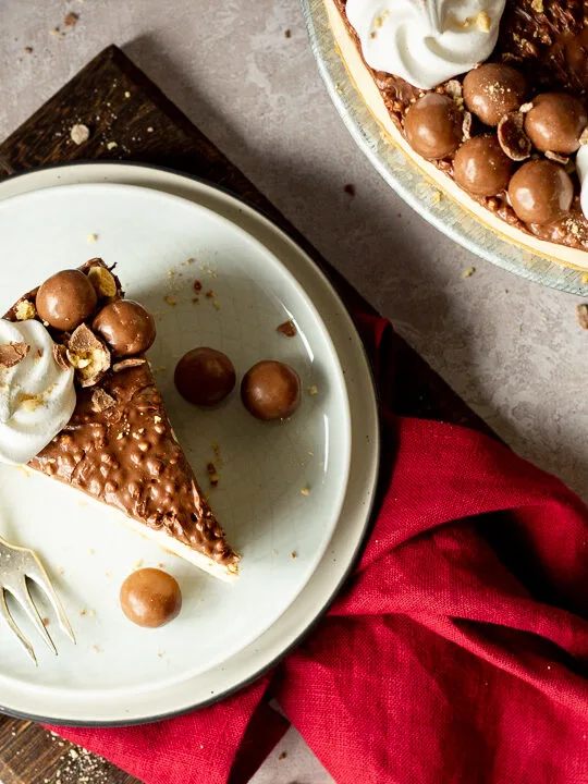 Birds eye view of a slice of no bake Malteser cheesecake on 2 stacked plates with a fork. There are crushed Maltesers scattered around, a red linen napkin and the remaining cheesecake on a cake stand in the background. Set on a light neutral backdrop.
