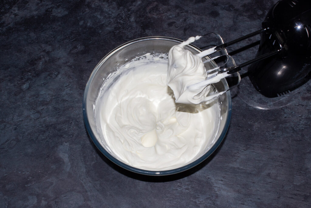 Double cream and icing sugar whipped to stiff peaks in a glass mixing bowl on a kitchen worktop.