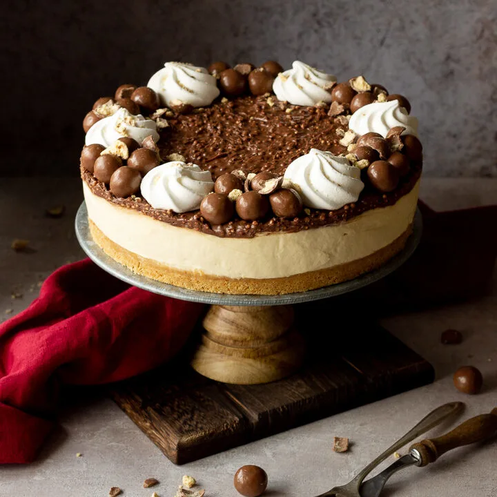 Front view of a no bake Malteser cheesecake on a cake stand. With crushed Maltesers scattered around, a red linen napkin and 2 forks. Set on a light neutral backdrop.