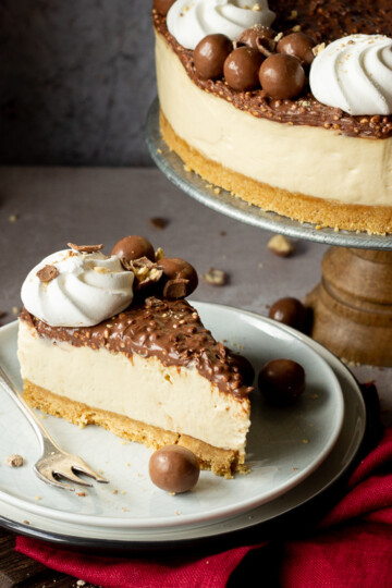 Front view of a slice of no bake Maltesers cheesecake set on 2 stacked plates with a fork. There are crushed Maltesers scattered around, a red linen napkin and the remaining cheesecake on a cake stand in the background. Set on a light neutral backdrop.