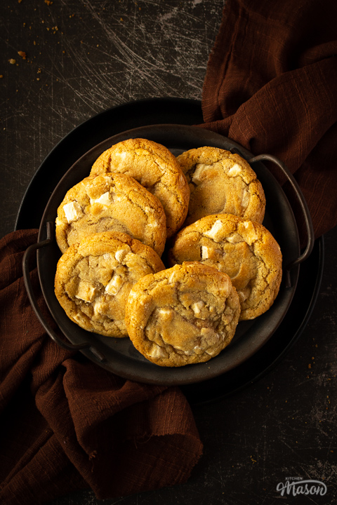 6 white chocolate chip cookies on an iron handled plate with a brown linen napkin underneath. Set on a scratched black metal backdrop.