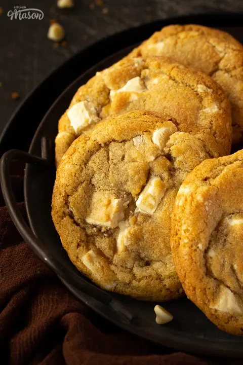 Close up of 4 white chocolate chip cookies on an iron handled plate with a brown linen napkin underneath. Set on a scratched black metal backdrop.