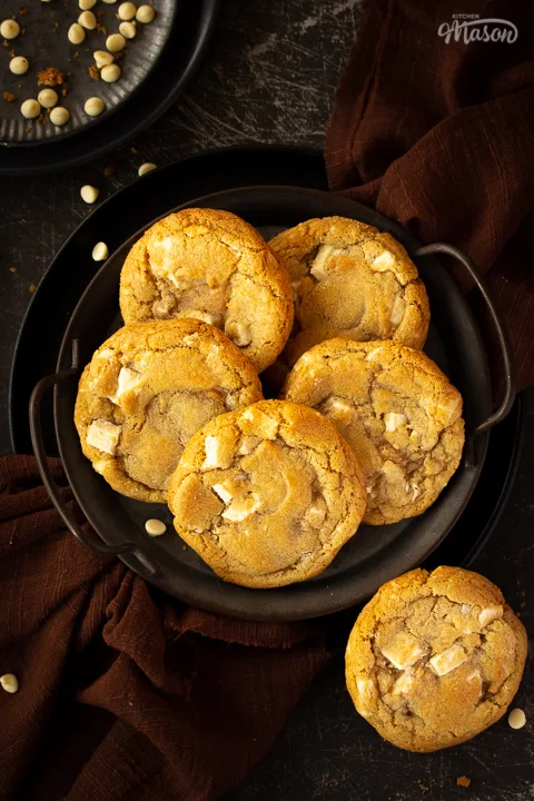 5 white chocolate chip cookies on an iron handled plate and 1 cookie to the side, with a brown linen napkin underneath. Set on a scratched black metal backdrop with white chocolate chips scattered around.