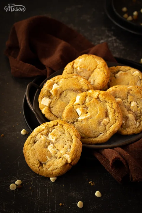 Front view of 5 white chocolate chip cookies on an iron handled plate and 1 cookie to the side, with a brown linen napkin underneath. Set on a scratched black metal backdrop with white chocolate chips scattered around.