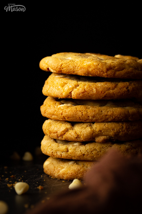 A stack of 6 white chocolate chip cookies set on a scratched black metal background with a brown linen napkin and white chocolate chips scattered around.