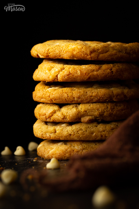 A stack of 6 white chocolate chip cookies set on a scratched black metal background with a brown linen napkin and white chocolate chips scattered around.