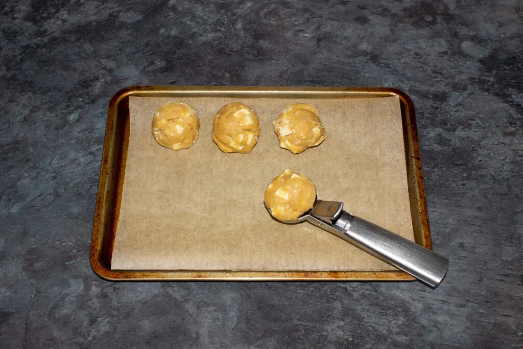 Balls of white chocolate chip cookie dough on a lined baking tray with an ice cream scoop