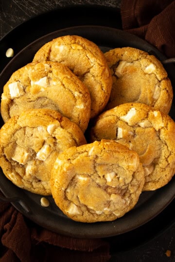 Close up of 6 white chocolate chip cookies on an iron handled plate with a brown linen napkin underneath. Set on a scratched black metal backdrop.