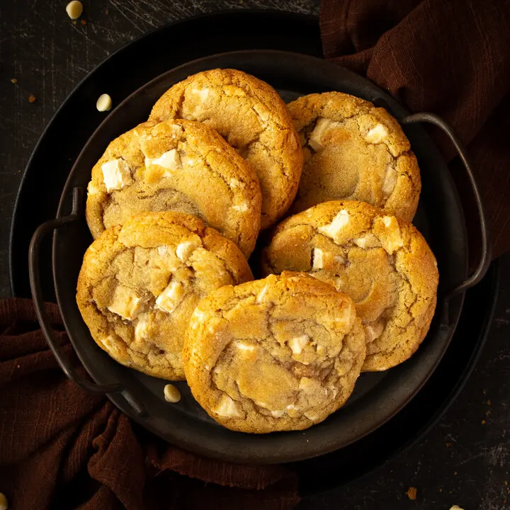 Close up of 6 white chocolate chip cookies on an iron handled plate with a brown linen napkin underneath. Set on a scratched black metal backdrop.