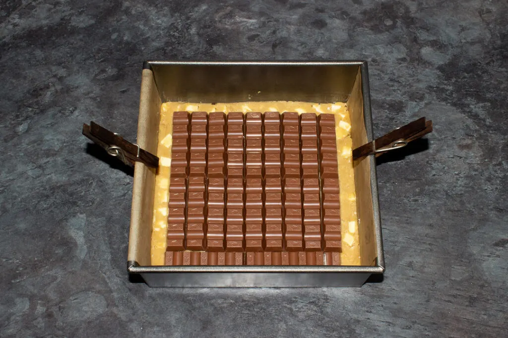 White chocolate chip cookie dough pressed into the bottom of a lined square baking tin, topped with lots of Kinder bars.
