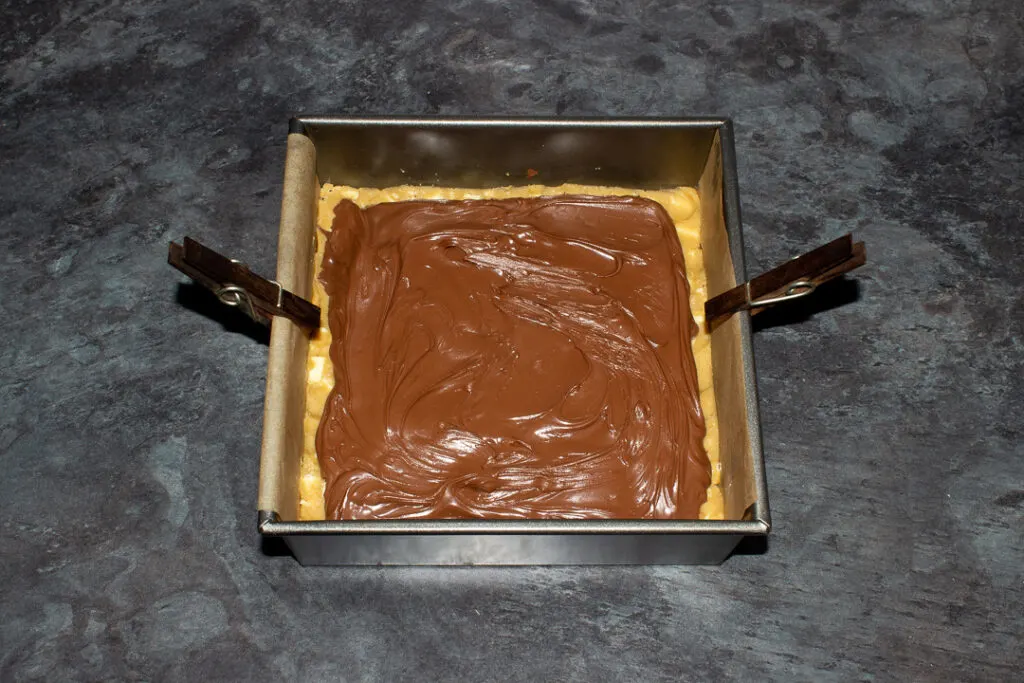 A lined square baking tin filled with 2 layers of white chocolate cookie dough, a layer of Kinder bars and a layer of Nutella spread.