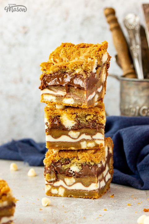 A stack of 3 white chocolate kinder nutella cookie bars on a cream backdrop. There are more bars in the background along with an iron tankard filled with cutlery and a blue linen napkin.