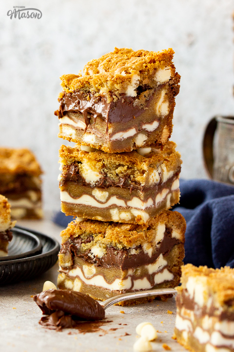 A stack of 3 white chocolate kinder nutella cookie bars on a cream backdrop. There are more bars in the background along with an iron tankard filled with cutlery, a spoon full of nutella and a blue linen napkin.