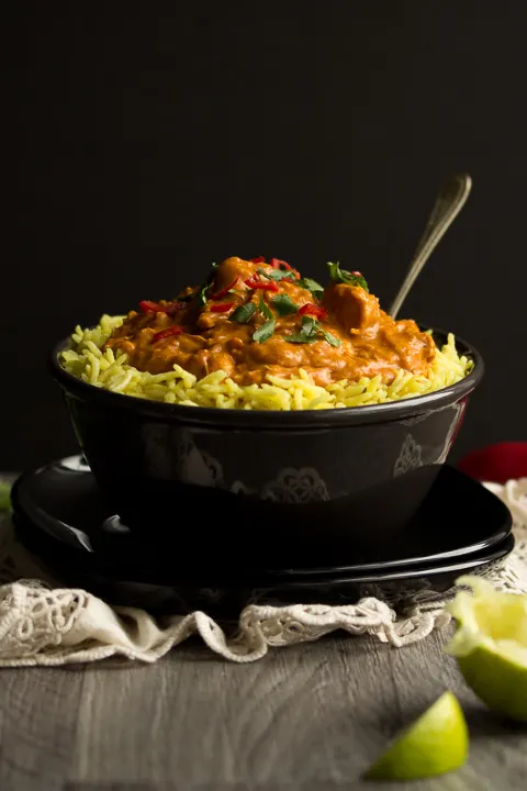 A side view shot of a black bowl filled with chicken curry over rice, topped with coriander and red chilli with a fork inside. There's a cream napkin and lime wedges in the background.