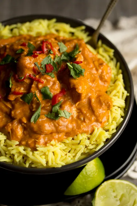 Close up overhead shot of chicken curry and rice in a black bowl on a black plate topped with coriander and chopped red chilli. Theres a spoon in the side of the bowl, a cream linen napkin and lime wedges in the background.