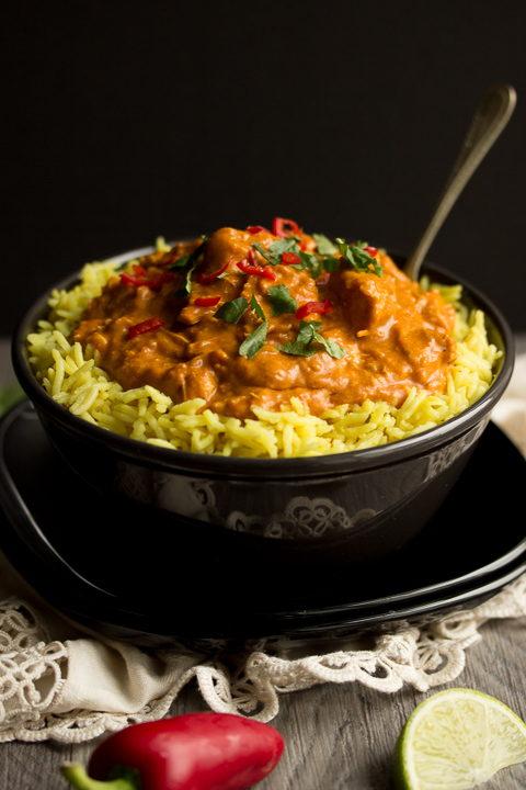 A black bowl filled with chicken curry over rice, topped with coriander and red chilli with a fork inside. Set on a black plate over a cream napkin. There's a whole red chilli and lime wedges in the background.