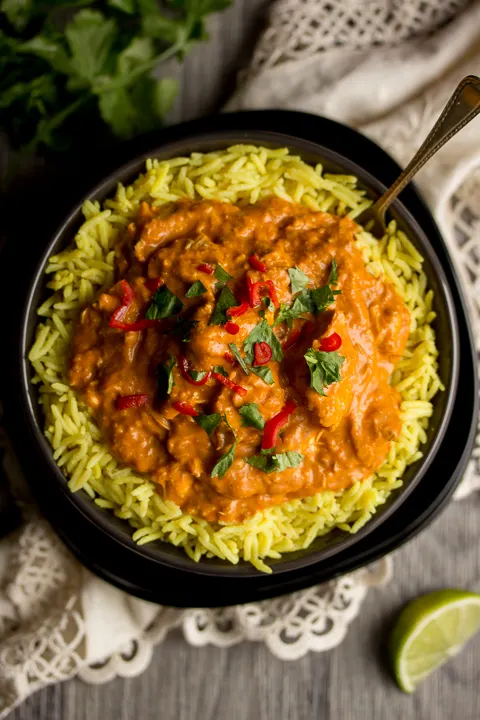 Birds eye view of chicken curry in a black bowl with rice topped with coriander and chopped red chilli and a spoon in the side. Set over a grey backdrop with coriander, lime wedges and a cream napkin in the background.