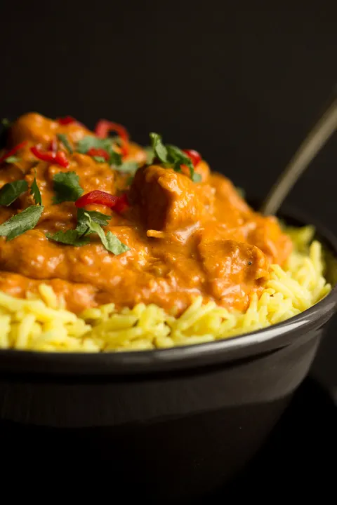 Close up of a black bowl filled with chicken curry over rice, topped with coriander and red chilli with a fork inside.