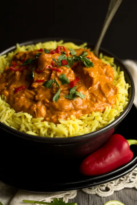 A black bowl filled with chicken curry over rice, topped with coriander and red chilli with a fork inside. Set on a black plate over a cream napkin with a whole red chilli in the background..