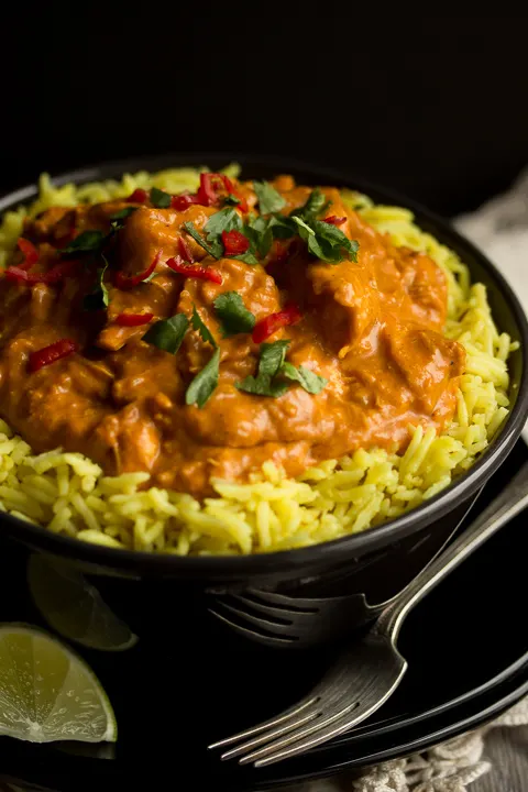 A black bowl filled with chicken curry over rice, topped with coriander and red chilli. There's a black plate underneath with a fork on it plus a lime wegde and a cream linen napkin in the background.