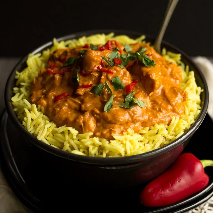 A black bowl filled with chicken curry over rice, topped with coriander and red chilli with a fork inside. Set on a black plate over a cream napkin with a whole red chilli in the background..