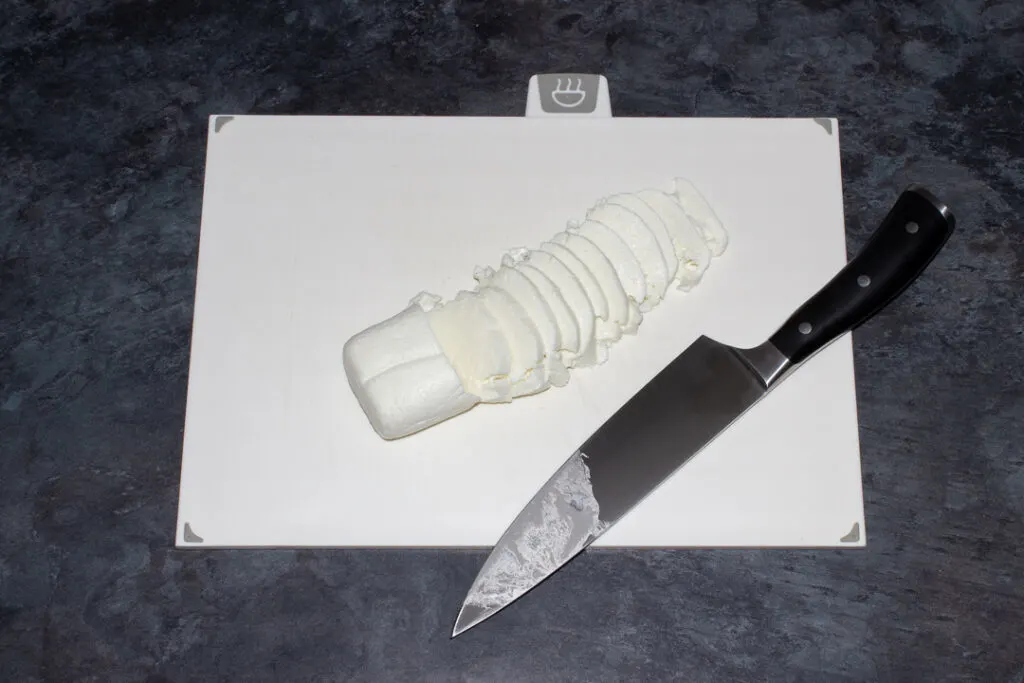 Mozzarella being sliced with a sharp knife on a white chopping board