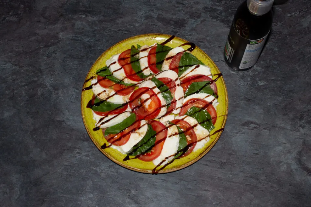 Slices of tomato, basil and mozzarella in a layered circle on a green plate with olive oil, balsamic vinegar and a balsamic glaze drizzled over the top.