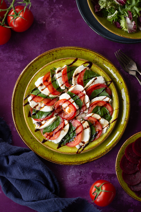 2 stacked green plates with a caprese salad drizzled with a balsamic glaze on top. Set on a purple backdrop, there is a bowl of lettuce, a plate of sliced beetroot, a blue linen napkin, 3 whole tomatoes and 2 forks in the background.