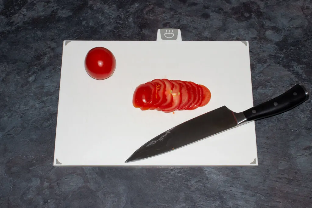a Tomato being thinly sliced with a sharp knife on a chopping board.