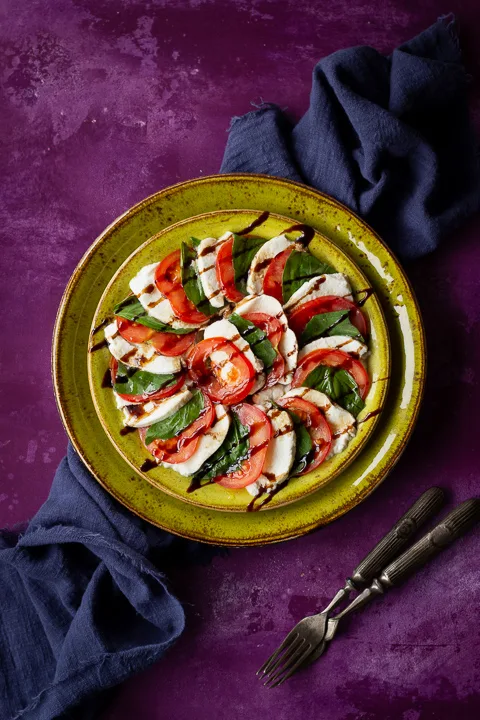 2 stacked green plates with a caprese salad drizzled with a balsamic glaze on top. Set on a purple backdrop there's also a blue linen napkin and 2 forks in the background.