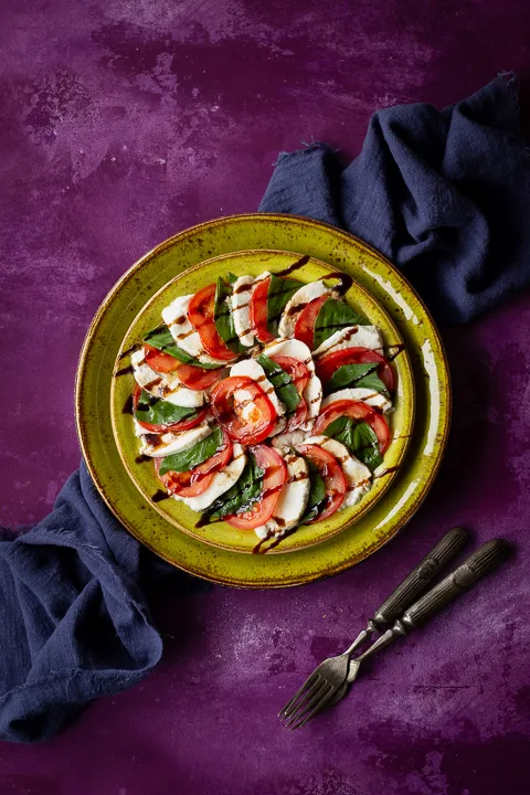 2 stacked green plates with a caprese salad drizzled with a balsamic glaze on top. Set on a purple backdrop there's also a blue linen napkin and 2 forks in the background.